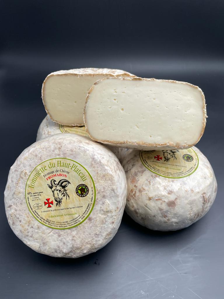 Demi-tomette chèvre à croûte naturelle, posée sur une pile de fromages, avec étiquette illustrée d'une tête de chèvre. Poids: 450g, lait de brebis thermisé.